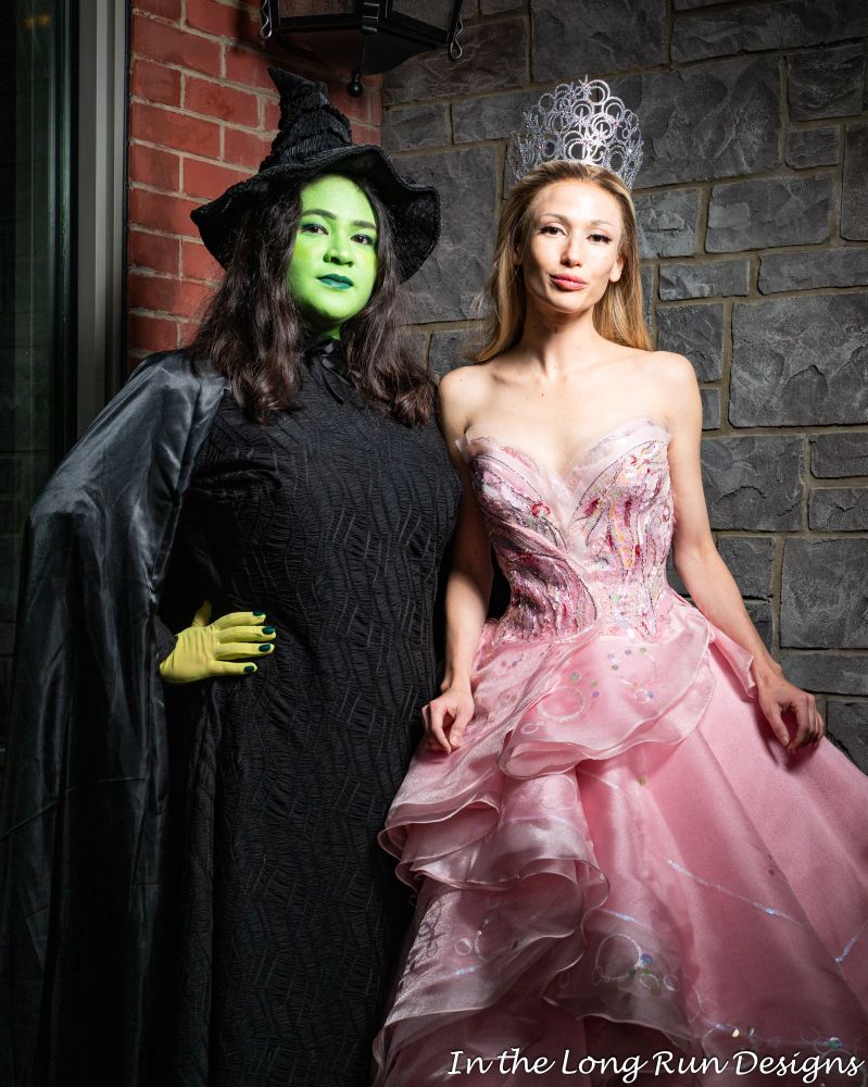 A three-quarter length photo of two cosplayers dressed as Elphaba and Glinda from Wicked (2024).