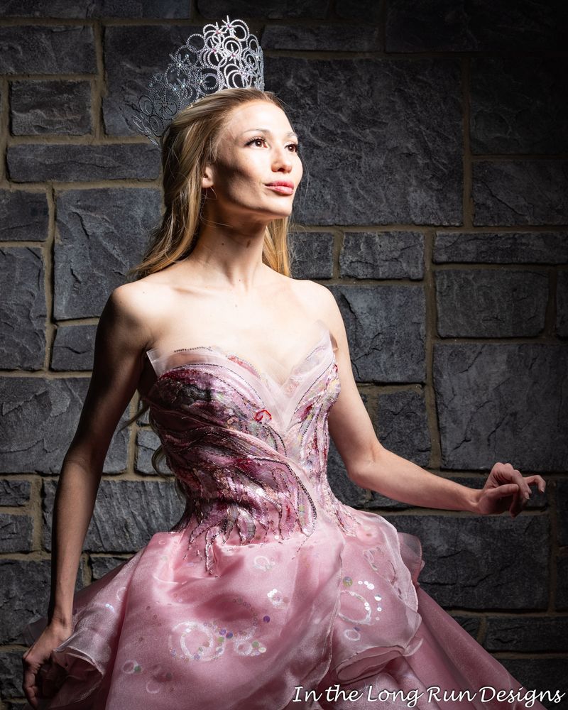 A half-length photo of a cosplayer dressed as Glinda from Wicked, standing in front of a stone wall. 