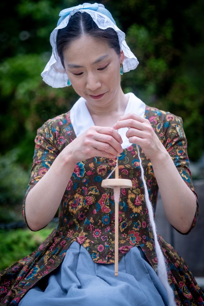 A woman in a 1780s style dress and white cap spinning wool with a drop spindle 