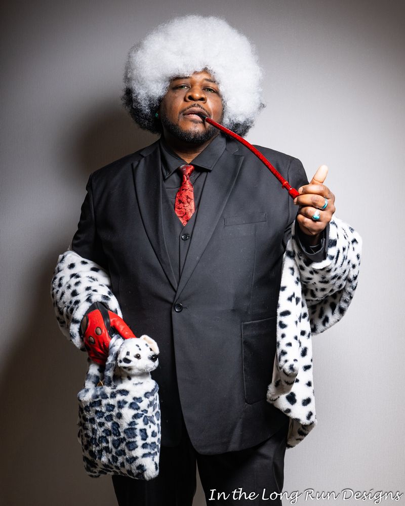 A three-quarter length photo of a cosplayer dressed as a masc version of Cruella de Vil. The costume includes a black suit, red tie, and Dalmatian print faux-fur stole. The cosplayer is smoking a long red pipe and holding a Dalmatian print bag containing a Dalmatian puppy plushy. 