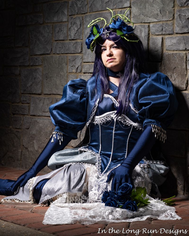A photo of a cosplayer dressed in a burlesque-inspired Mistress 9 costume from Sailor Moon, seated on a brick floor against a stone wall. 
