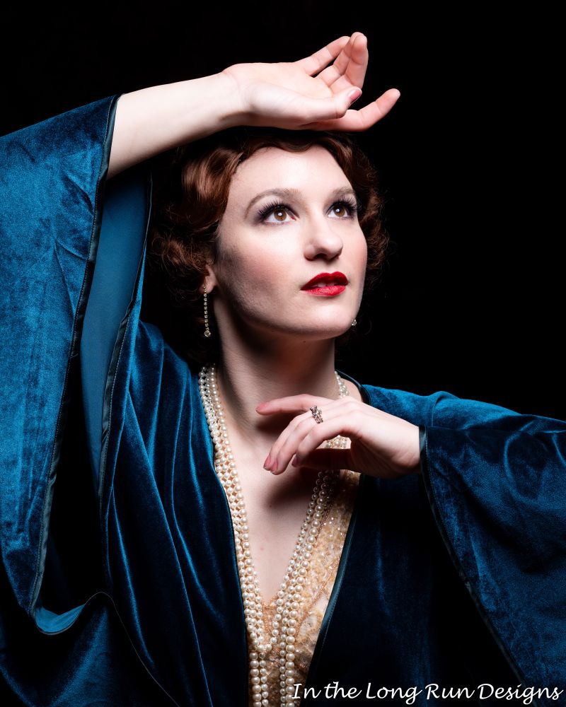 A quarter-length photo of a cosplayer dressed as a silent film star in a blue robe wearing a string of pearls.