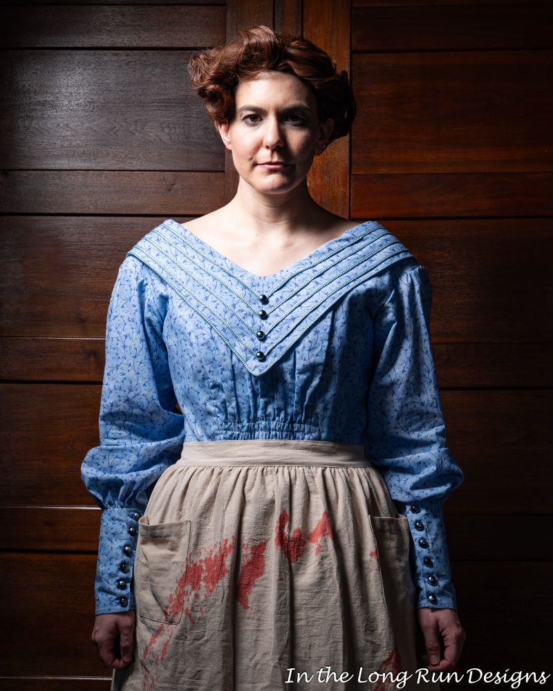 A three-quarter length photo of a cosplayer dressed as Mrs. Lovett from Sweeney Todd, wearing a blue dress and a white, blood-stained apron. The cosplayer stands in front of a dark wood-paneling background. 