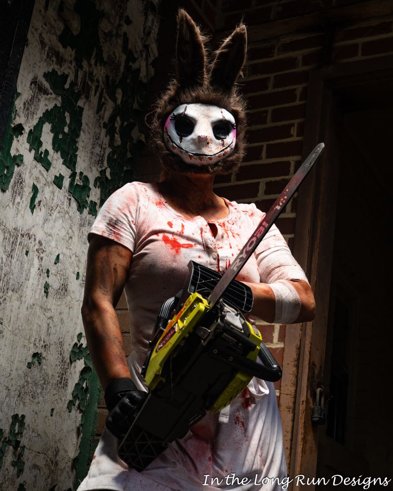 A three-quarter length photo of a cosplayer dressed in a horror costume, consisting of a bloodstained white T-shirt and apron, and a white animal mask with furry ears. The cosplayer is welding a chainsaw at the camera. 