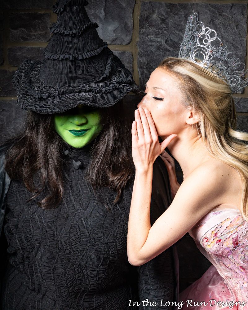 A half-length shot of two cosplayers dressed as Elphaba and Glinda from Wicked (2024), recreating the original Broadway poster for Wicked. The brim of Elphaba’s pointy hat shadows her eyes, leaving only her smile showing, while Glinda leans in and whispers in her ear. 