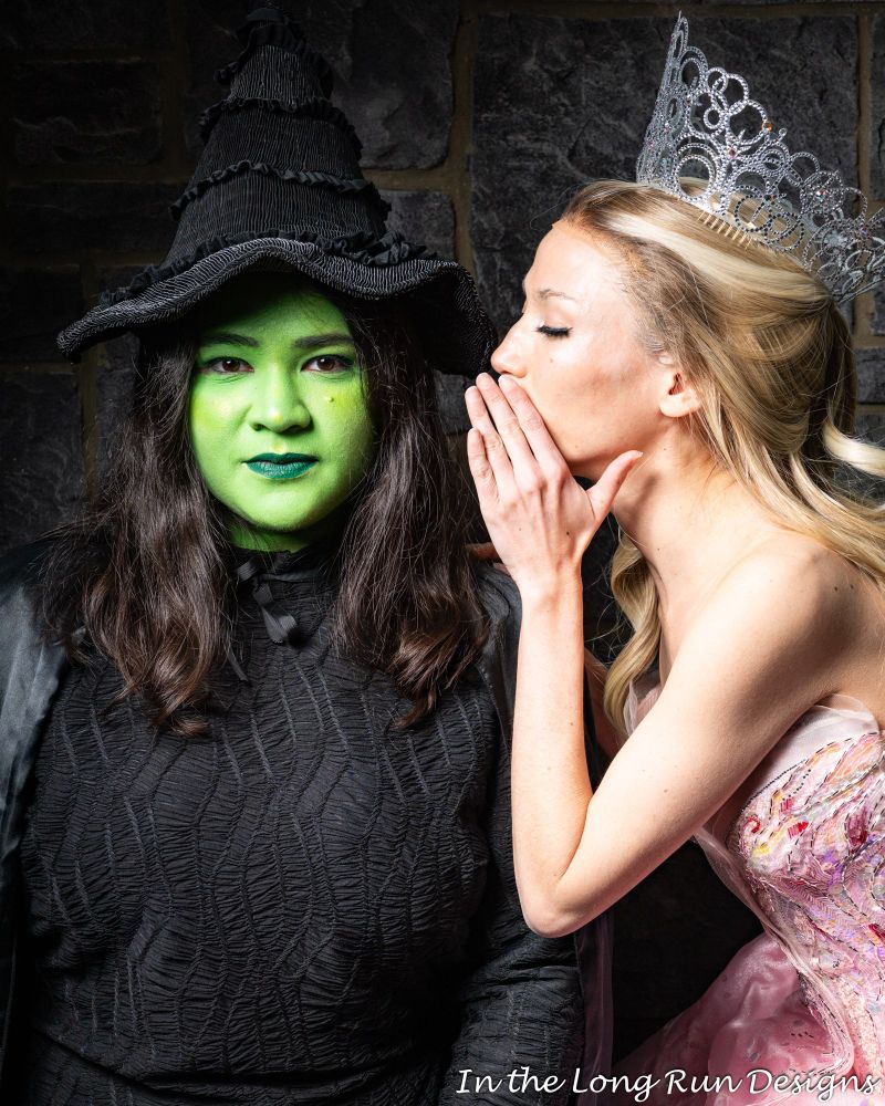 A half-length shot of two cosplayers dressed as Elphaba and Glinda from Wicked (2024), recreating the film’s poster. Elphaba stares straight at the camera with a neutral expression, while Glinda leans in and whispers in her ear. 