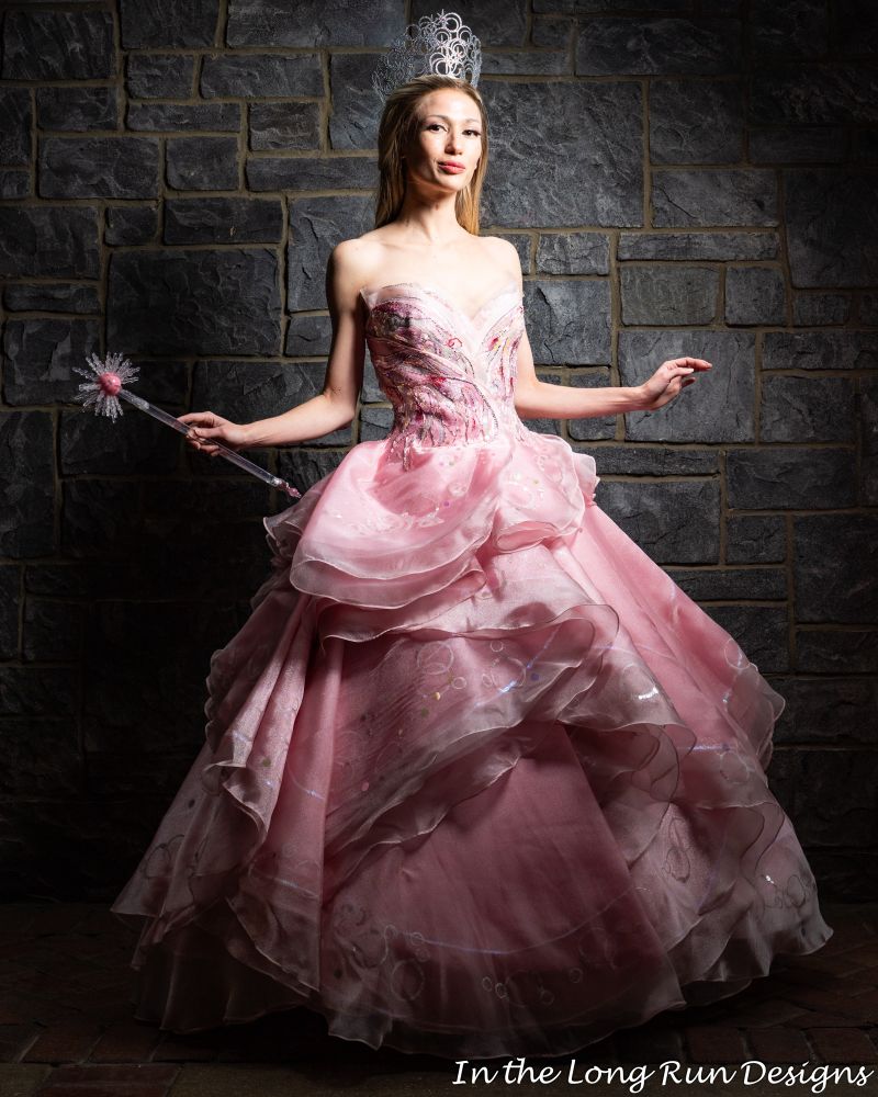 A full length photo of a cosplayer dressed as Glinda from Wicked, standing in front of a stone wall. 