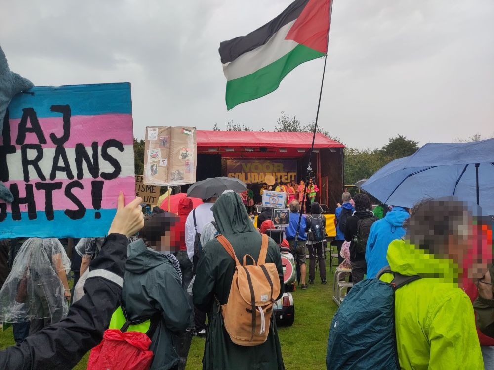 Foto van een stoet mensen voor een podium. Zo'n 20 mensen zijn in beeld. Links is een protestbord in de kleuren van de trans vlag. In het midden wappert iemand de vlag van Palestina. Op het podium hangt een banner met "voor solidariteit".