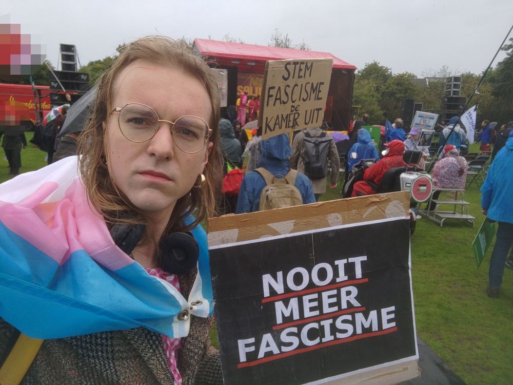 Selfie. Ik hou een bord vast met de tekst "nooit meer fascisme" en ik heb een trans vlag om mijn schouders. Ik sta op een grasveld waar het regent en ik kijk serieus in de camera. Achter mij is een groep mensen te zien die kijkt naar een podium. Een persoon heeft een protestbord met de tekst "stem fascisme de kamer uit"