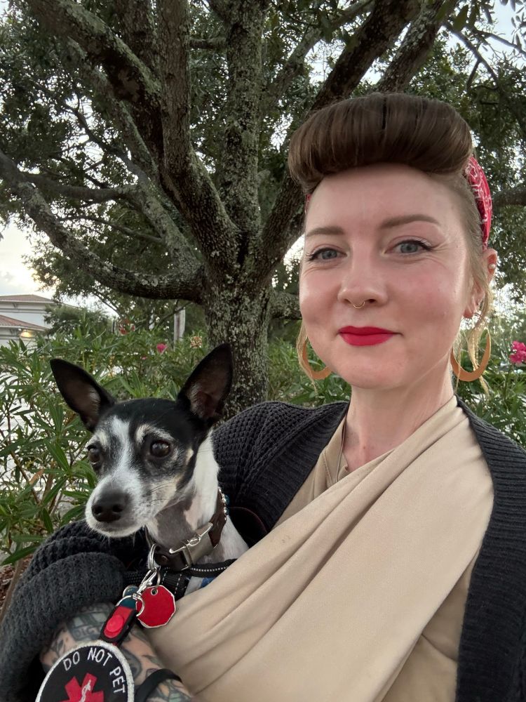 a white femme person stands outside in front of a tree. they have red lipstick on and a small cats eye for makeup. their bangs are rolled into a bumper and their hair is pulled up behind that, with a red bandana tied around everything. they’re holding a rat terrier wearing a do not pet collar sign. they’re smiling. the terrier looks perpetually worried 