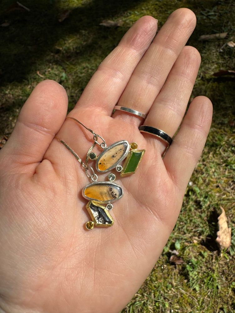 a white right hand holds two pendants  with grass in the background. the pendants have oval agates as the main stone, clear with shades of brown and black speckles. a smattering of yellow and light green sapphires are tube set around the pendants. below each agate is an angular stone: one pendant is green jade, the other is a clear quartz with black striped rutilations within it