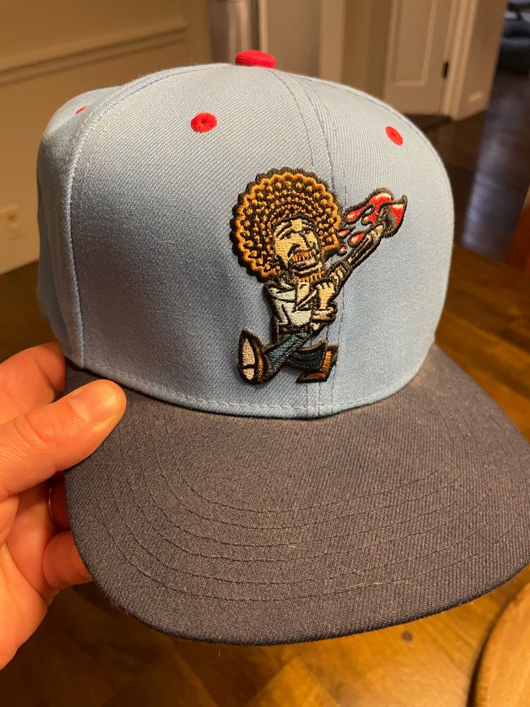 Blue baseball cap with a cartoon Bob Ross swinging paint brushes like a baseball bat