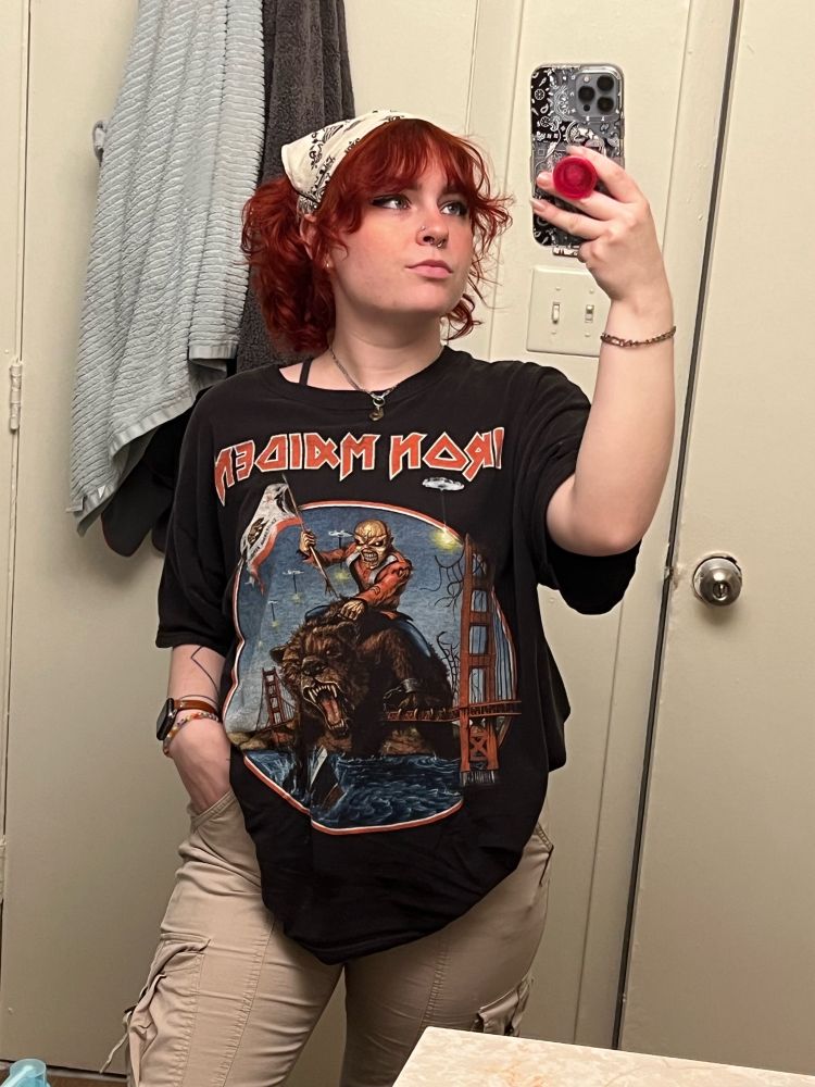a mirror selfie of me, a curly redhead with bangs in an oversized iron maiden tour t-shirt and tan cargo pants. the top of my curls are hidden beneath a tan bandanna with black patterning. my right hand, which has my apple watch and a rainbow hair tie on its wrist, is tucked into my pocket while my left hand holds my iphone, assisted by a hot pink popsocket on a bandanna patterned phone case