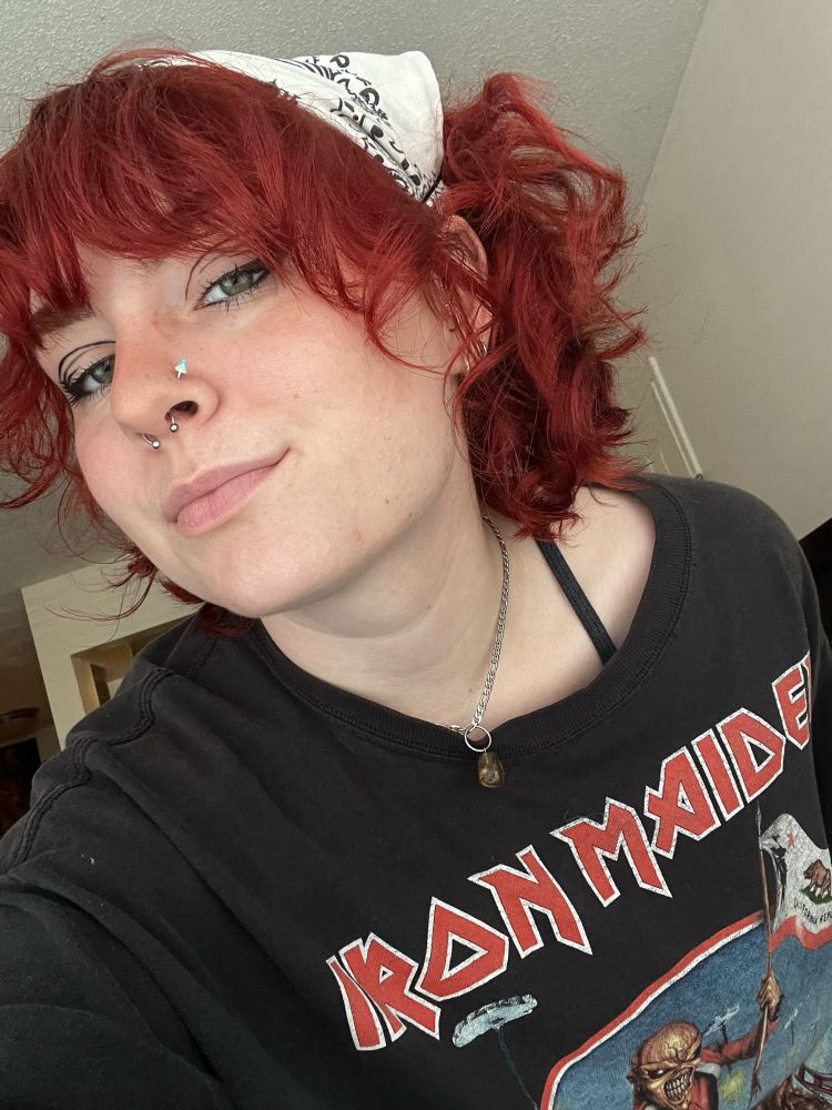a front-camera selfie, showing off a bit of my graphic eyeliner, my septum ring, and my nostril piercing (which is a blue, green, and white tie dye cone). my short chain necklace is dangling down, showing my citrine pendant. them curls be poppin 