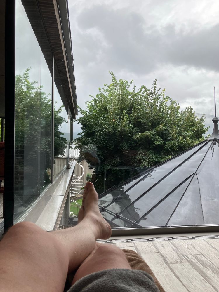 & wieder Pims nackte Beine, diesmal auf einer Indoorpoolliege mit Blick durch eine Glasfront auf ein verregnetes Blechdach & eine Linde, darüber sich grade auflösende Regenwolken.