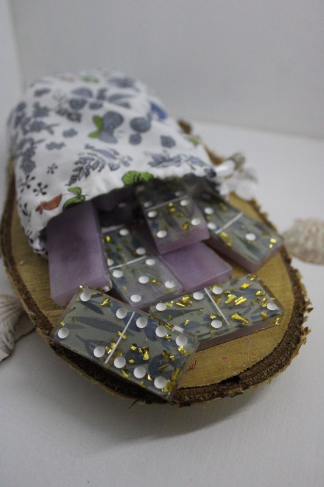 Photo d'un jeu de dominos. Celui-ci a été réalisé en résine transparente. On peut voir quelques tuiles de dominos, posées sur un rondin de bois, et sortant d'une pochette en tissu. Les tuiles ont le dos violet, et la face à pois fleurie dans des tons violets. Les pois sont peints en blanc. 
Une pochette de rangement permet de ranger le jeu, celle-ci est blanche avec des fleurs bleues-violettes et a été cousue à la machine