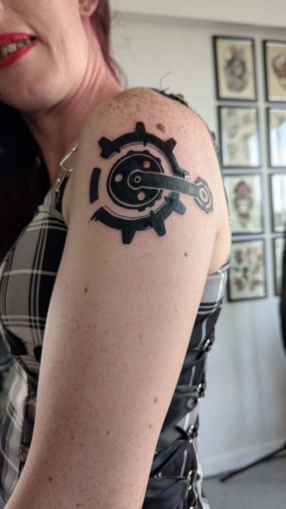 A fresh tattoo imitating Vi's neck tattoo in Arcane, on a woman's shoulder. She is wearing a dark chequered dress with ribbon lacing and red lipstick. 