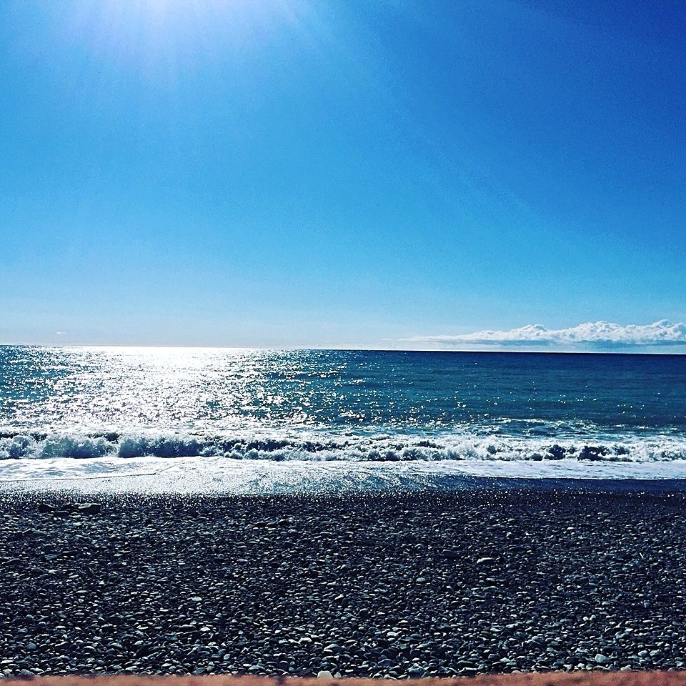 Vy över stenstrand mot blått hav mot horisonten med vågskum mot stranden, en klarblå himmel och solstrålar som glittrar i vattnet. 
