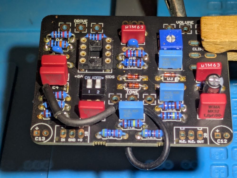 A small (about 2 inches on each side), nearly square, black printed circuit board with numerous capacitors, resistors, and other components (mostly blue or red in color) attached, suspended horizontally about 1/2 inch above a blue work surface by a wooden clothespin in the upper right corner. A black wire attached to one leg of a red capacitor winds from left to right across the frame and disappears below the board near the front center.