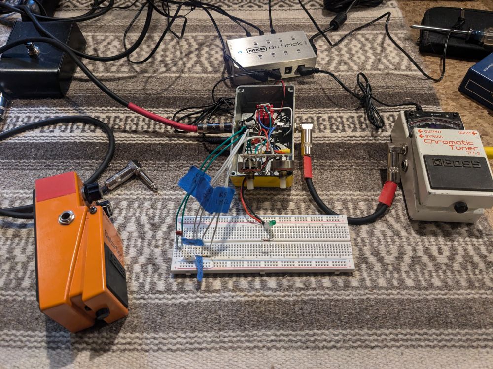 An overturned DOD 250 overdrive pedal on a rug, with wires connecting it to a breadboard with various electronic components stuck into it. On the left is a Boss DS-1 pedal on its side, and on the right is a Boss TU-2 tuner.