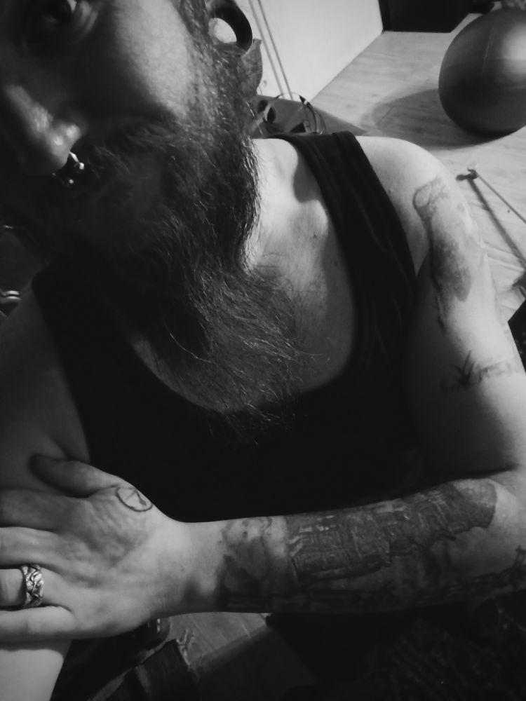 Schwarz weiß Foto einer bärtigen Person in einem Tanktop gekleidet mit Septum und einem Vegan V auf der Hand. Der linke Arm ist parallel über die Brust verschränkt 