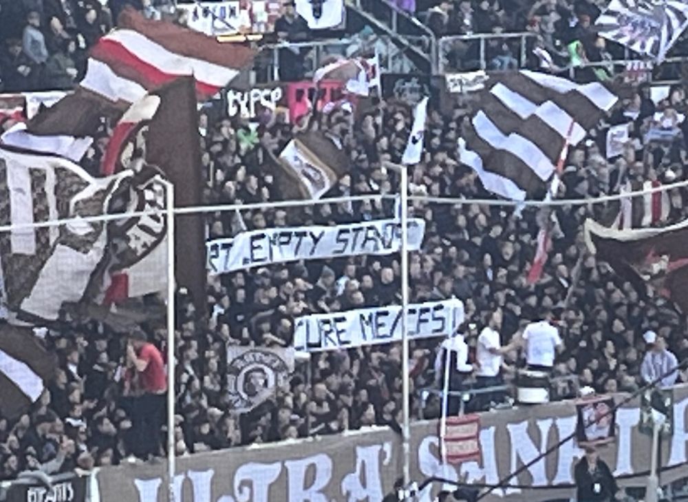 Auf zwei Tapeten in einer vollen Stehplatzkurve beim FC St. Pauli steht „Support „Empty Stands““
„Cure ME/CFS“.
Das Wort „Support“ ist auf dem Bild von einer Fahne verdeckt. 