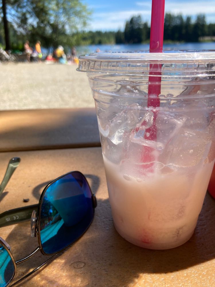 Italian soda and sunglasses on a picnic table with sandy beach and water in the distance