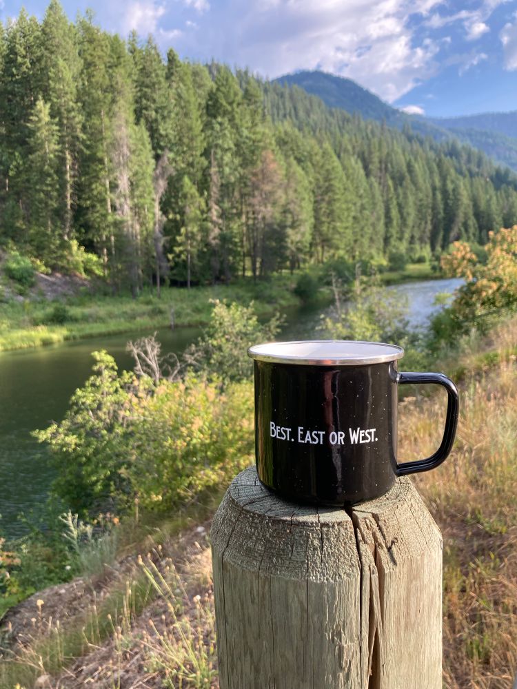 Tin cup sitting on a fence post with the phrase, “Best. East or West” printed on it. River, trees and mountains in the background.