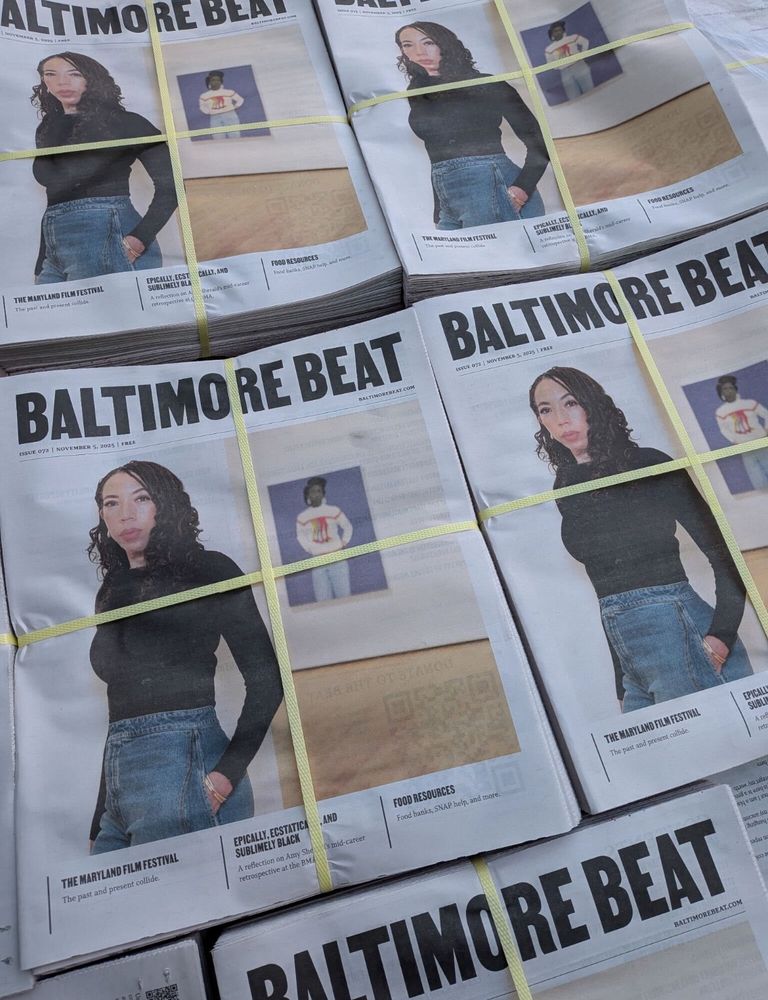 Stacks of copies of Baltimore Beat. The cover features a photo of artist Amy Sherald. She wears a black top and blue jeans. 