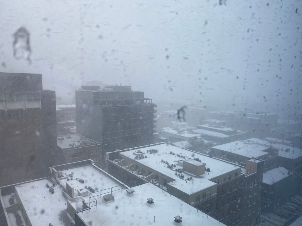 A photo taken from a high-rise showing snow-covered city rooftops obscured by ice pellets seeping down the windowpane and active blustery snowfall. 