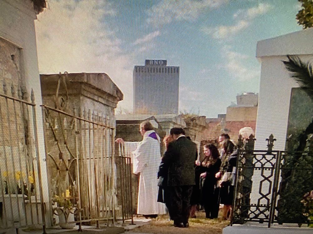 A photo of a computer monitor showing a movie scene, a group of mourners in a historic New Orleans cemetery full of above-ground crypts. In the background, a single brutalist office building looms. 