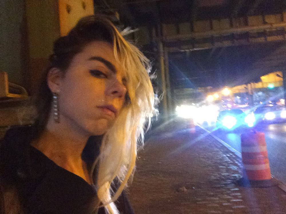 A white woman with bottle blonde swept-over hair scowls at the viewer standing on grimy Third Avenue beneath the BQE. The glare of oncoming traffic fills the right side of the frame.