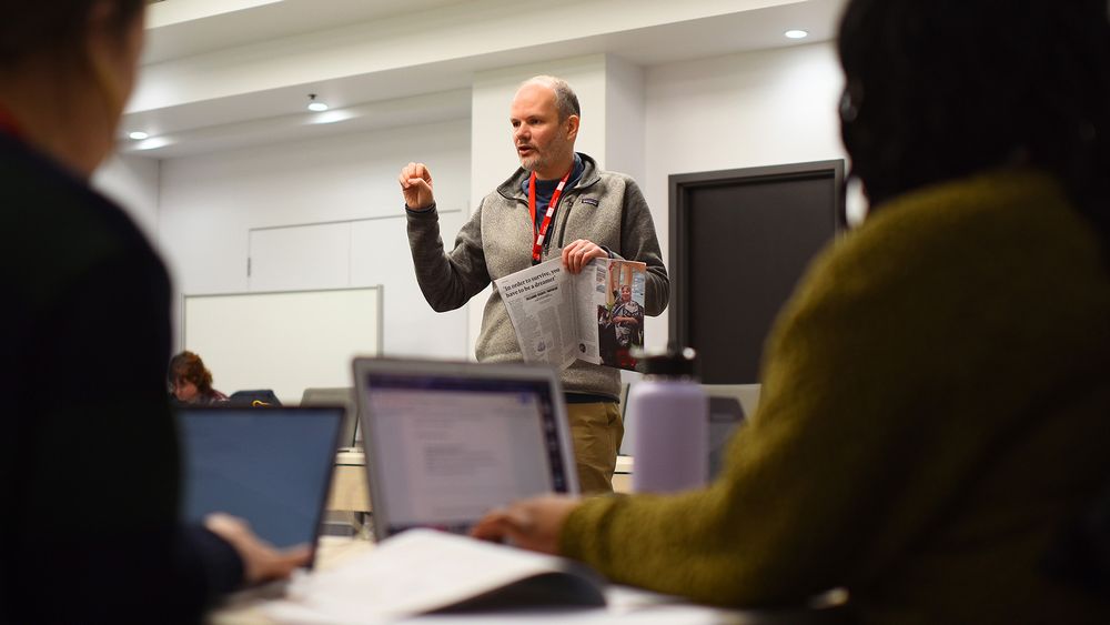 A man stands and speaks to a group in a classroom while holding papers; two people in the foreground are seated with laptops and notebooks.