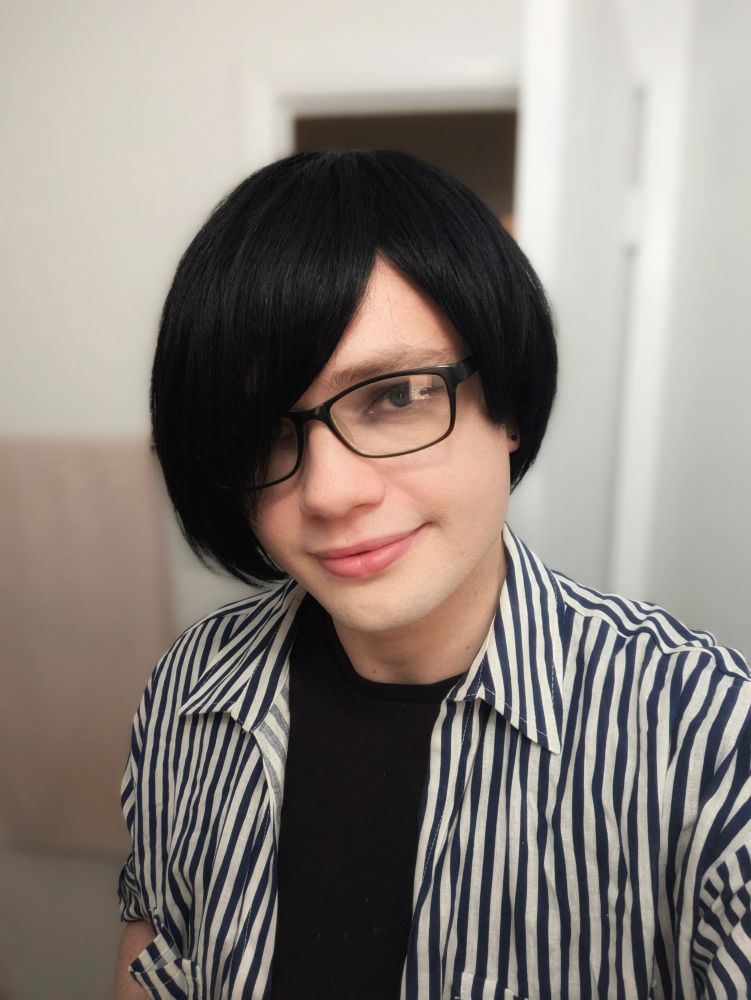 A selfie photo of me in my bathroom. My hair is black and my long, asymmetrical bangs cover my right eye. My left earlobe is exposed and I have a black stud earring. I'm wearing a short-sleeve collared shirt with narrow vertical white and navy stripes over top of a black T-shirt.