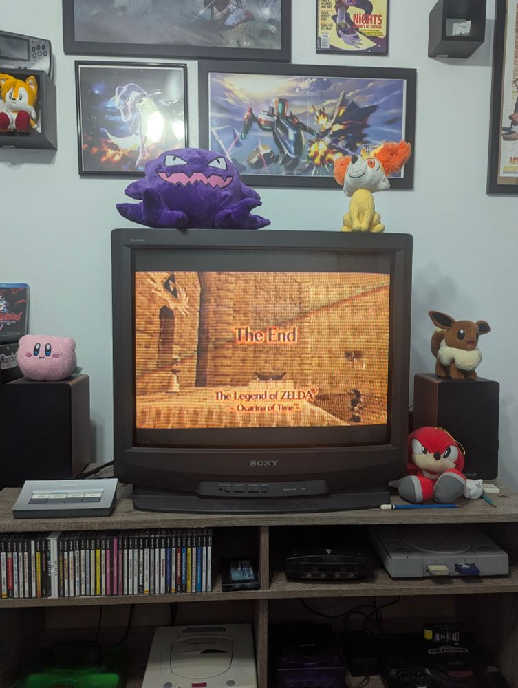 Same as the prior photo, but this time the television displays the final shot of the game after the credits. Text reading "The End - The Legend of ZELDA - Ocarina of Time" is superimposed over a sepia-tone view of an early-game cutscene in which Link and Zelda meet each other for the first time as children in the Hyrule Castle garden.