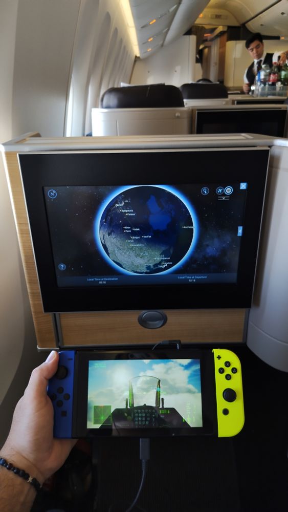 My view from seat 12A on the main Business class cabin of a Swiss Intl Air Lines Boeing 777-300ER. The middle third of the image shows the 15" 16:9 screen of the entertainment system, framed in black plastic and displaying the Earth globe in the middle atop a space background. The flight route is shown, stretching from Japan over the Arctic to Zurich. The screen sits on a hardshell console with a plywood finish. The upper third is a blurred view into the cabin, drenched in a warm white light and lined with beige walls and dark brown leather seatbacks. The lower third shows me holding a Nintendo Switch console with my left hand. The Joycon controller on the left is blue, the one on the right is neon green. The screen shows Ace Combat 7 running on the system, displaying an F-16 cockpit in a light green tint. 