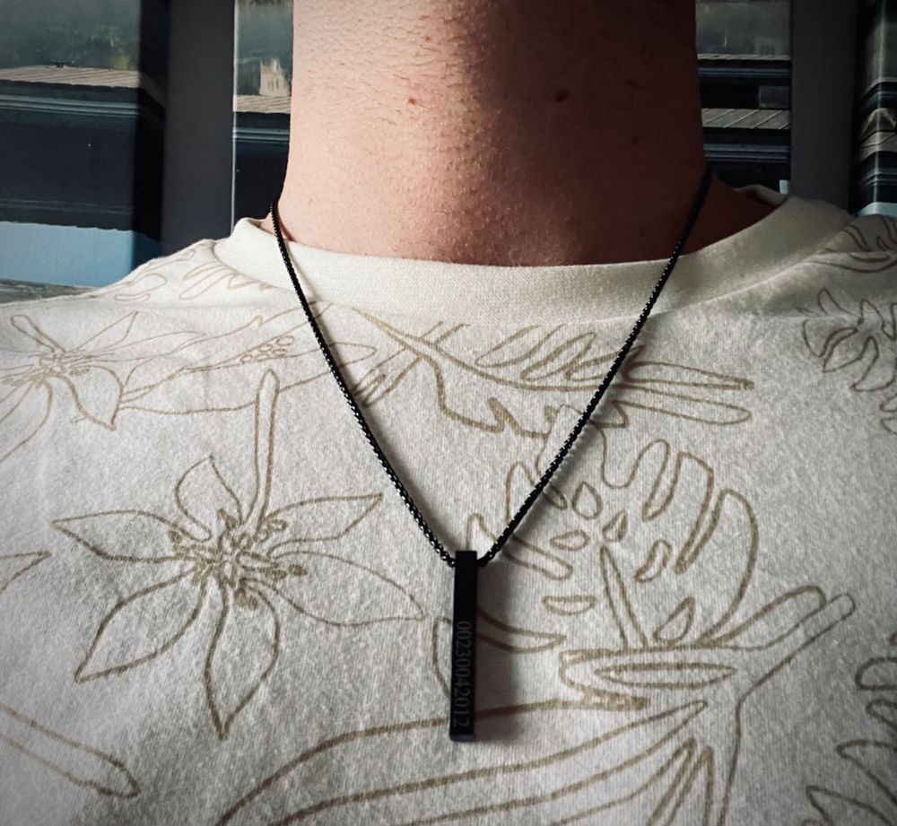 Pendant with engraved numbers around a submissive male’s neck, wearing a floral print tee shirt.