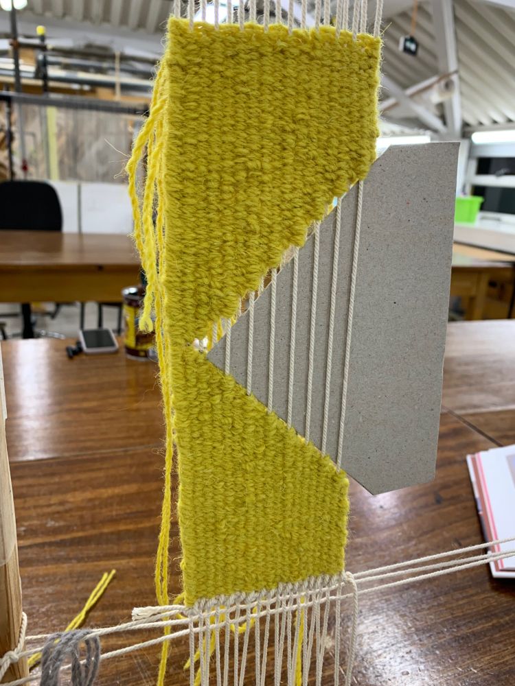 A photo of the tapestry sample on the frame. The tapestry is made with bright yellow wool and has a large triangle of cardboard sat in the warps to allow the tapestry to be woven with no warp in that area. The yellow wool of the tapestry, from top to bottom, makes a 45 degree slope from the right hand side to the left, and then a 45 degree slope from left to right. Those two 45 degree angles are important for the final 90 degree turn in the sample ;)