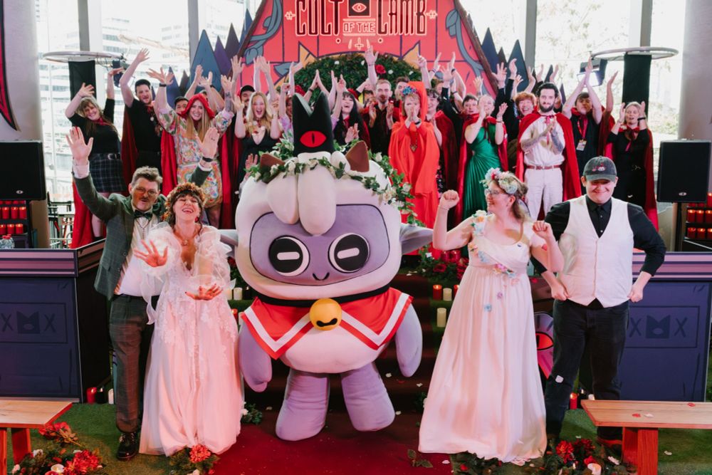 Two newly married couples stand beside a Lamb mascot from Cult of the Lamb, in front of a custom-made temple and a bunch of their friends and family. If these people haven't been deeply affected by art, I don't know who has!