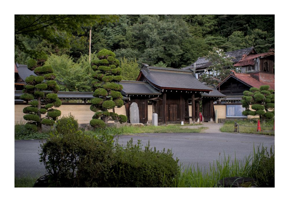 The outside of a shrine in Takayama City in Gifu, Japan. The entrance to the shrine is the main subject and it is made up of 3 doors, only the right side one is open. There are two tall bushes on the left of the shrine door, a stone tablet with some Kanji written on it, and more buildings to the right of it. A low bush peeks out in the foreground.
