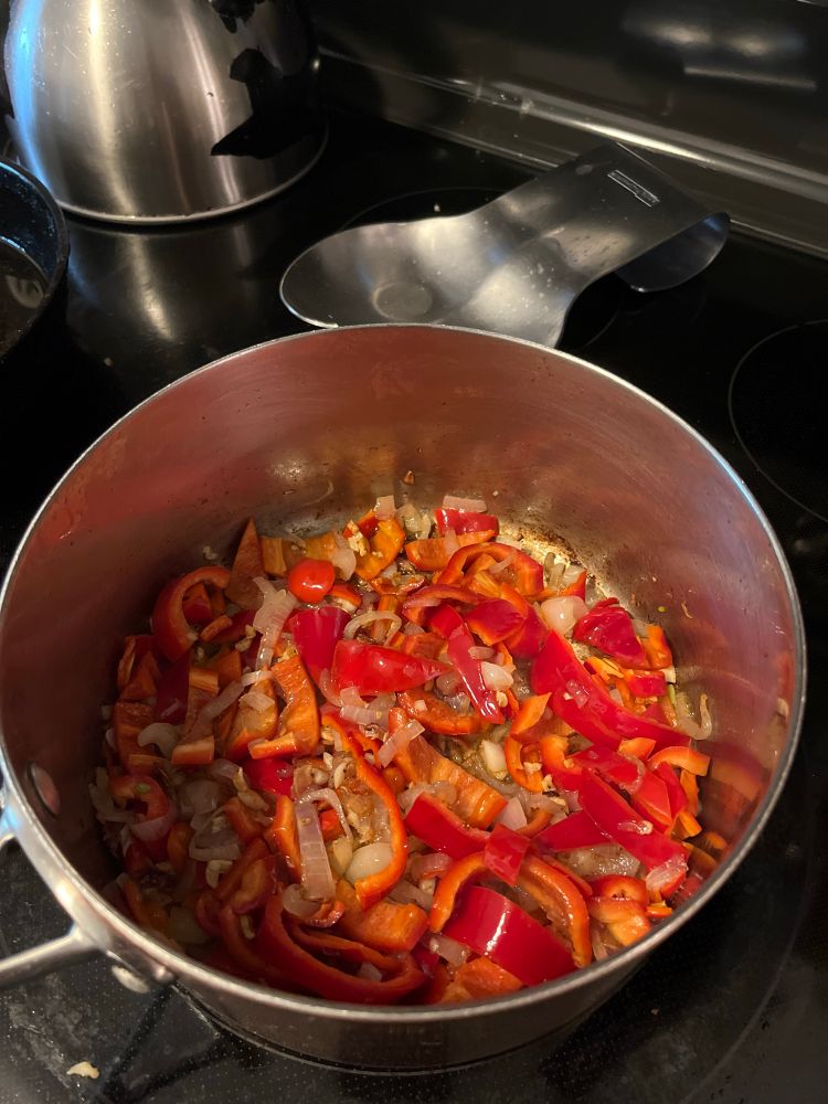 A copper saucepan on a black stove has chopped bright red peppers cooking in oil with minced shallots and garlic.