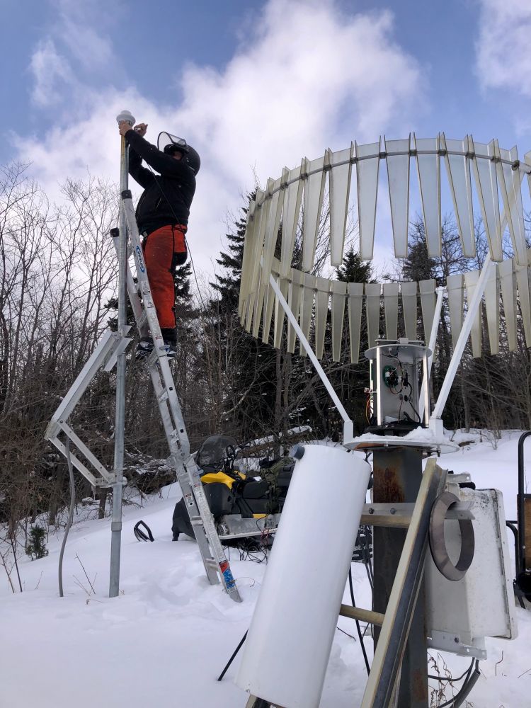 Person on a ladder next to a dismantled rain gauge, snow on the ground and snowmobile in background 