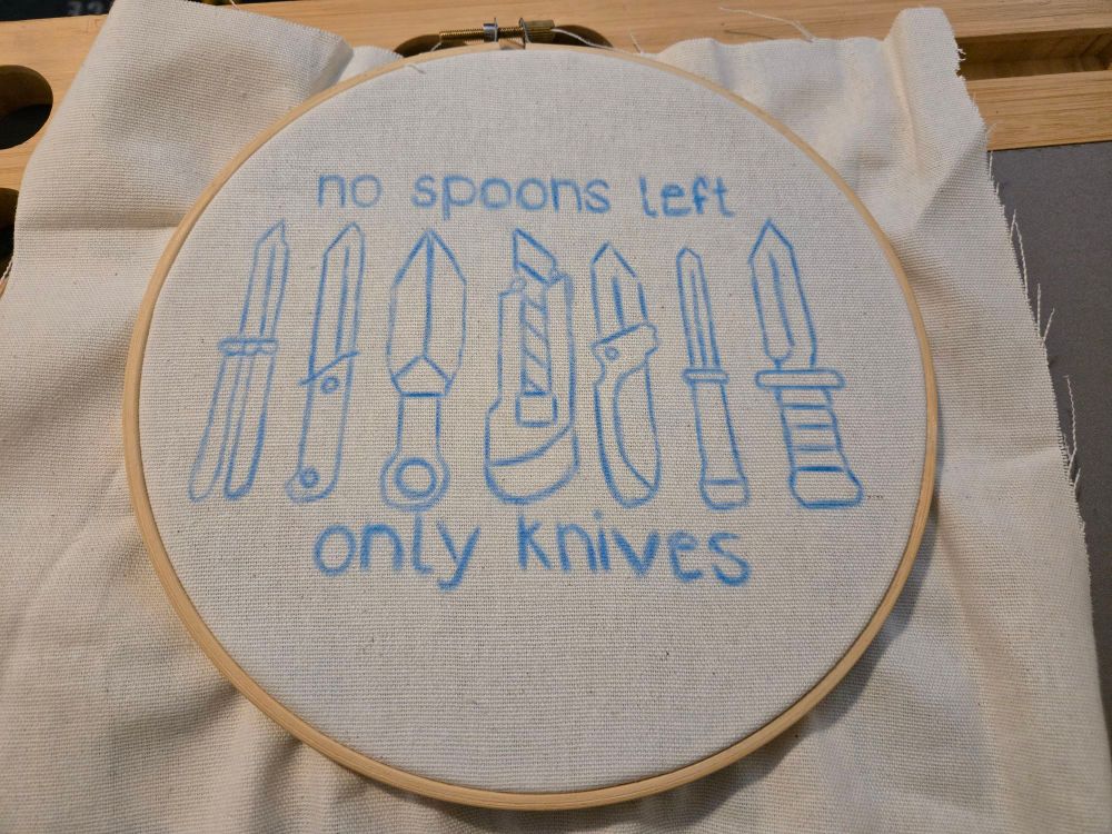 A pattern on an embroidery hoop. It features 7 different knives with the words "no spoons left only knives " written around them. The pattern is drawn in blue washable pencil. 