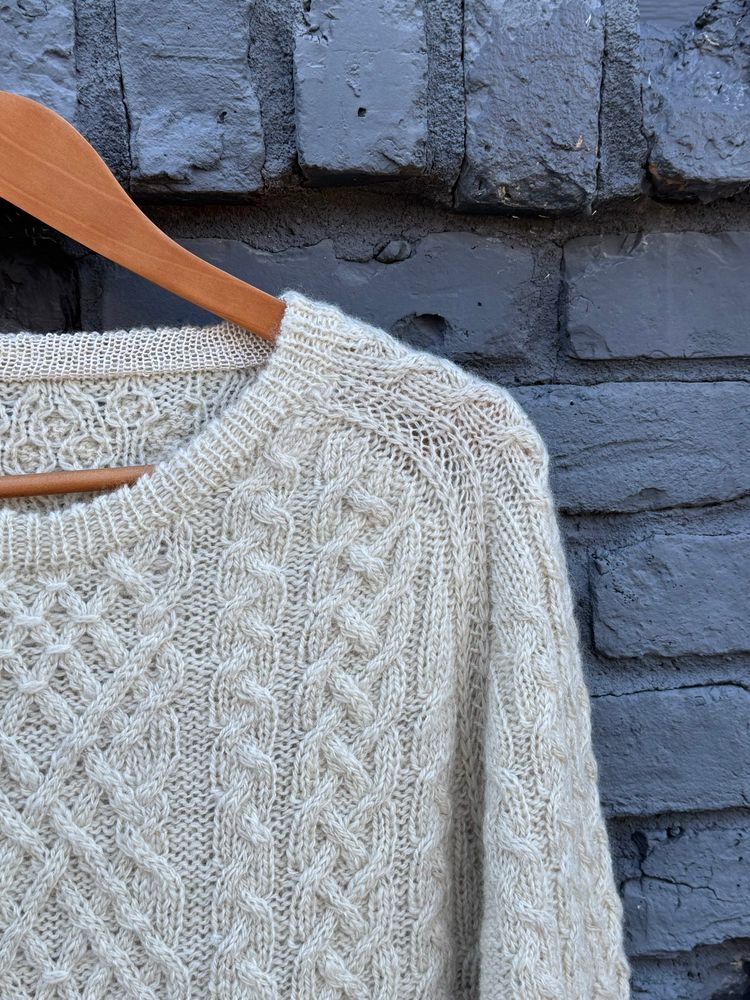 Close up of a cream colored, heavily cabled sweater hanging in front of a grey brick wall. This focuses on the collar and left sleeve. The inside of the neckband is trimmed in silk.