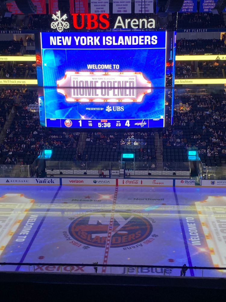 Ice rink with the Islanders’s logo (white NY on top of an orange geographical image of Long Island, all over a blue background with an orange border). The screen above reads “welcome to the home opener” and shows the score of 4-1, Capitals currently winning. The top of the screen says UBS Arena