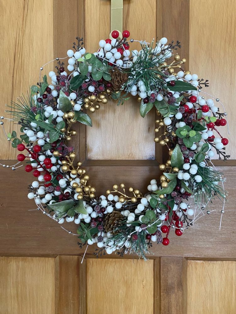 A Christmas wreath, with berries of white, bright red, deep red, and gold, with various green foliage and 2 little pinecones. There are some silver beads threaded through that poke out so it’s a little messy and rustic looking. It is hanging on a wreath hanger on an unpainted wooden door.