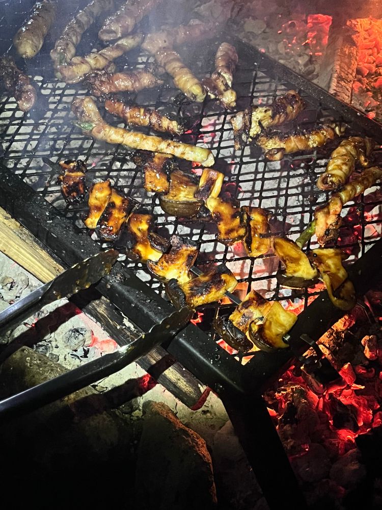 A campfire grill lit by flashlight, showing skewers of marinated mushrooms, bacon wrapped enoki, and bacon wrapped asparagus cooking over hot coals 