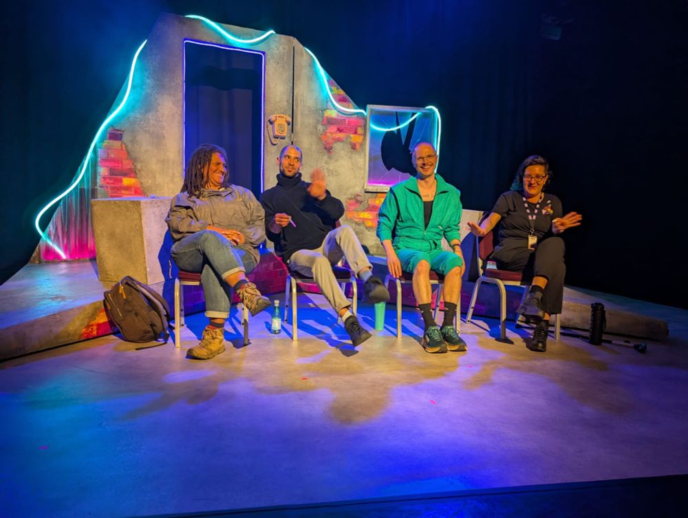 A group of people sat on stage smiling and laughing in front of a colourful, brightly lit stage set with pink and grey brickwork. 