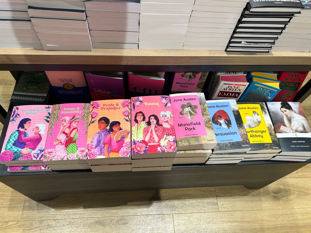 A row of different Jane Austen novels in a book store. The left ones are pink ones with modern colorful drawings of couples, the right ones are more subdued in color and style. 