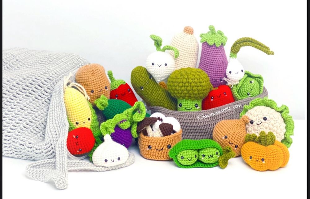 an assortment of crochet amigurumi vegetables with a white background. some of them are in a grey bag, while others are in a darker grey basket. each vegetable has two small black eyes and a smiley face, making the veggies look cute and comforting